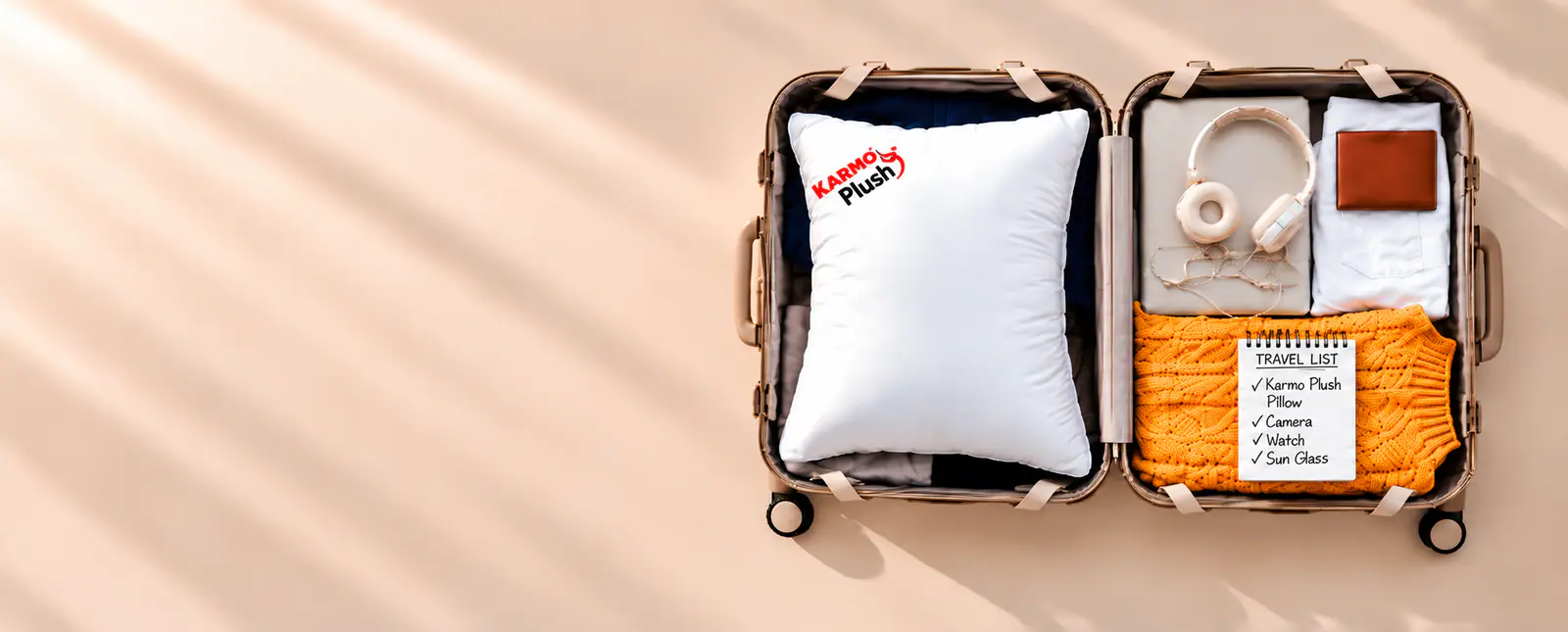 A Karmo Plush pillow packed in an open suitcase beside headphones, a wallet, an orange knit and notepad, on a warm cream backdrop